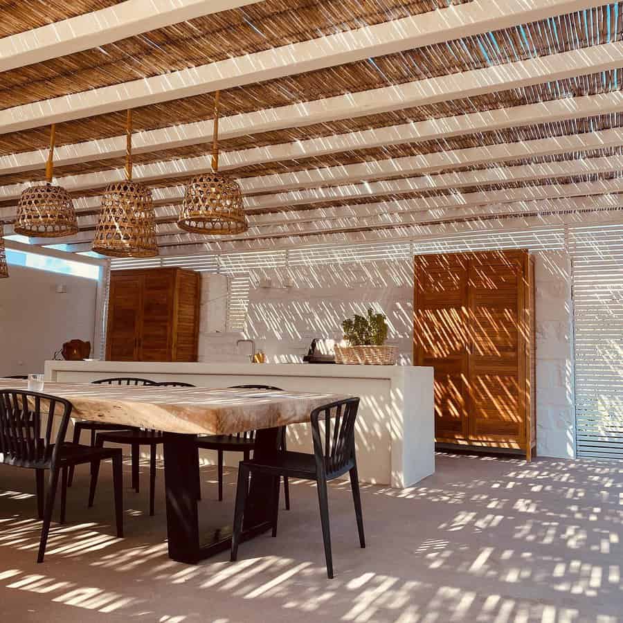 Outdoor dining area with wooden table, black chairs, wicker pendant lights, and slatted roof casting shadows on white walls