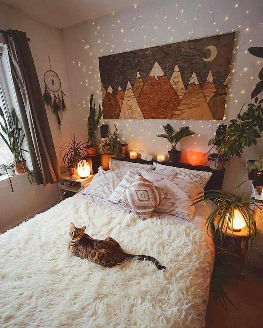 Boho bedroom with a fluffy bedspread, a cat lounging on the bed, string lights, plants, and a mountain-themed tapestry on the wall