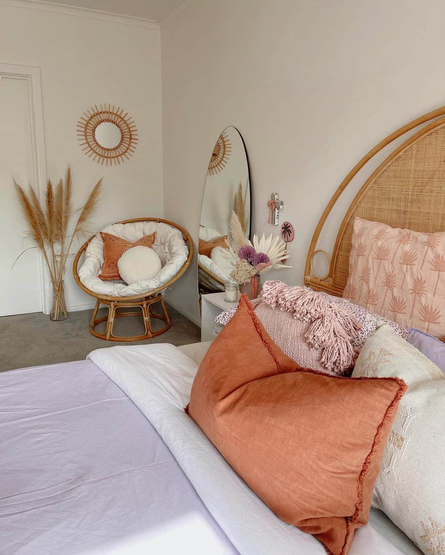 Bedroom with a cozy wicker chair, a large mirror, decorated with sunburst art, bed adorned with orange and white pillows and pampas grass