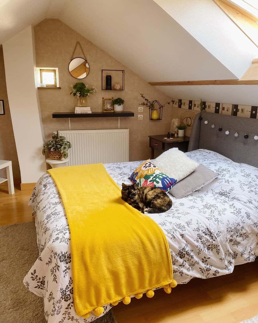 Cozy attic bedroom with warm neutrals, a mustard throw, and boho decor. Soft lighting, plants, and a relaxed cat add charm to the space.