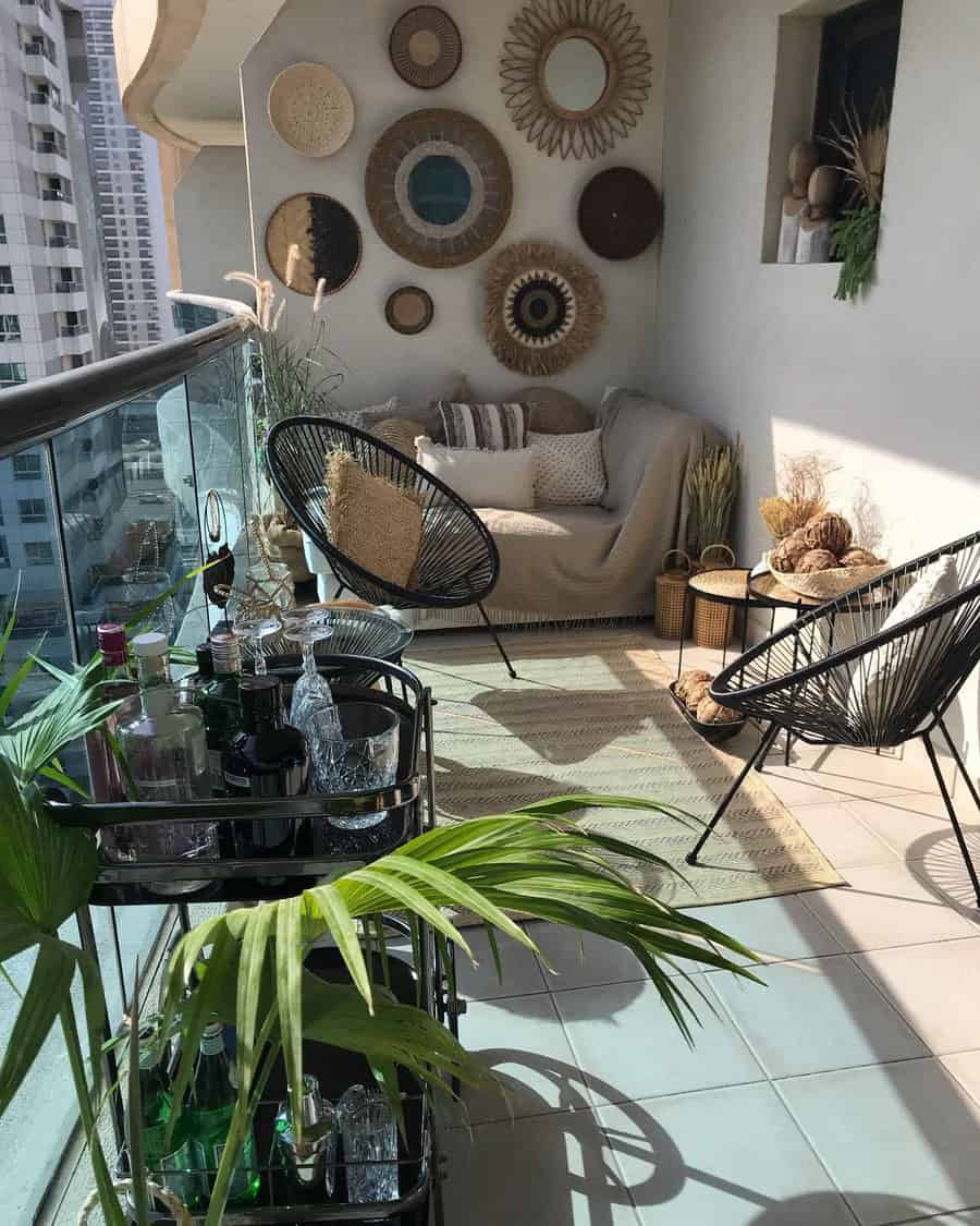 A cozy balcony with a sofa, wicker wall decor, two black chairs, plants, and a glass bar cart with bottles and glasses, offering an urban high-rise view