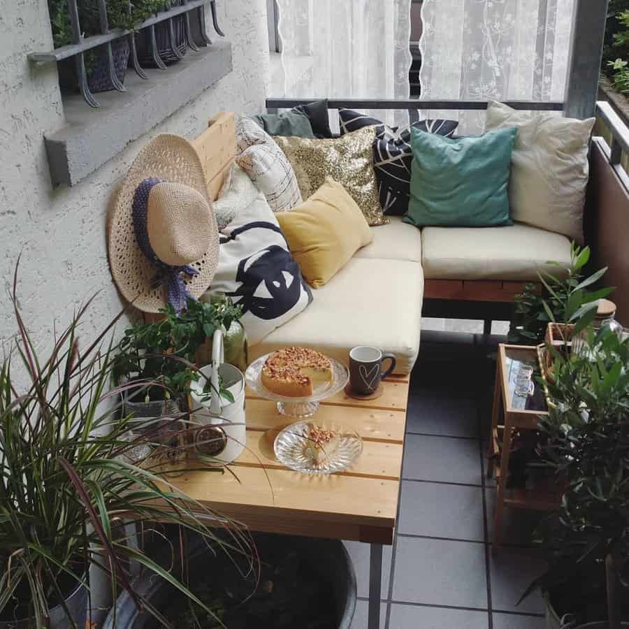 Cozy boho balcony with a corner sofa, colorful cushions, lush greenery, and a wooden table set for coffee and cake, creating a relaxing nook