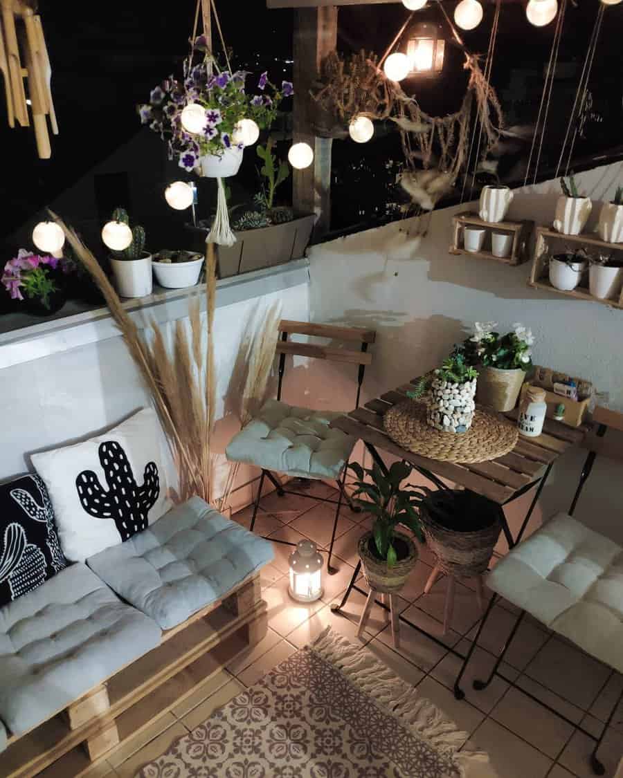 Boho-style balcony at night with string lights, a wooden table, cushioned pallet seating, potted plants, and hanging decor