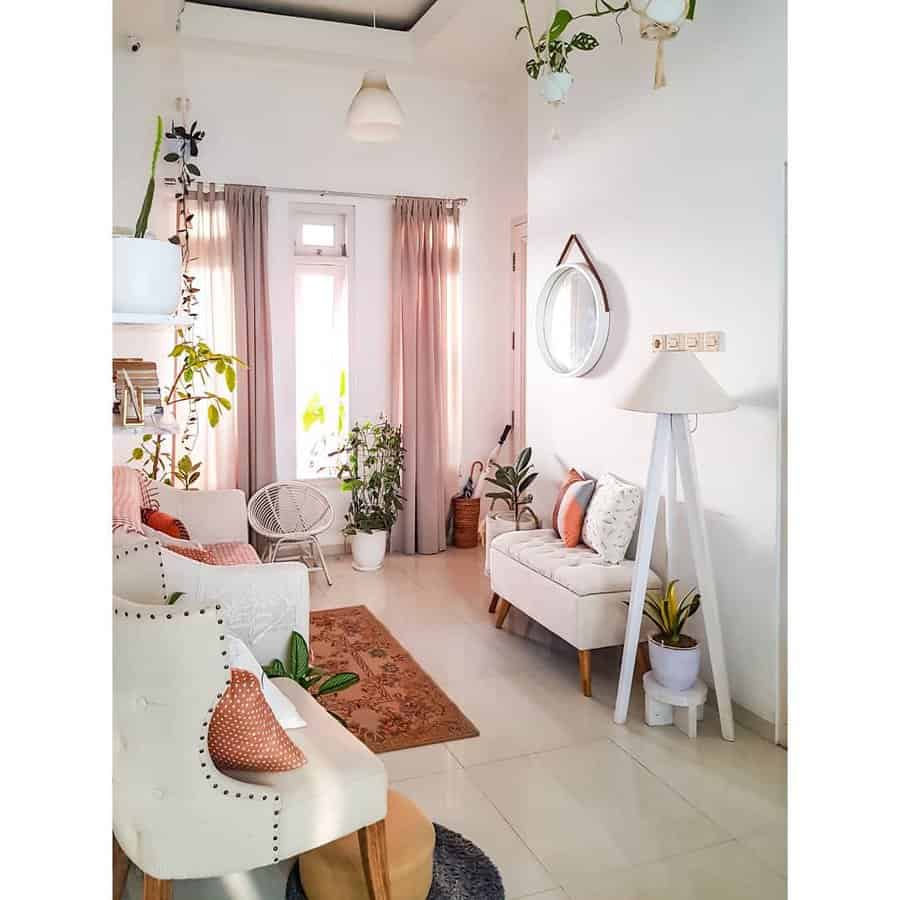 Bright living room with white furniture, green plants, beige curtains, and a rug; wall decor includes a round mirror and hanging pots