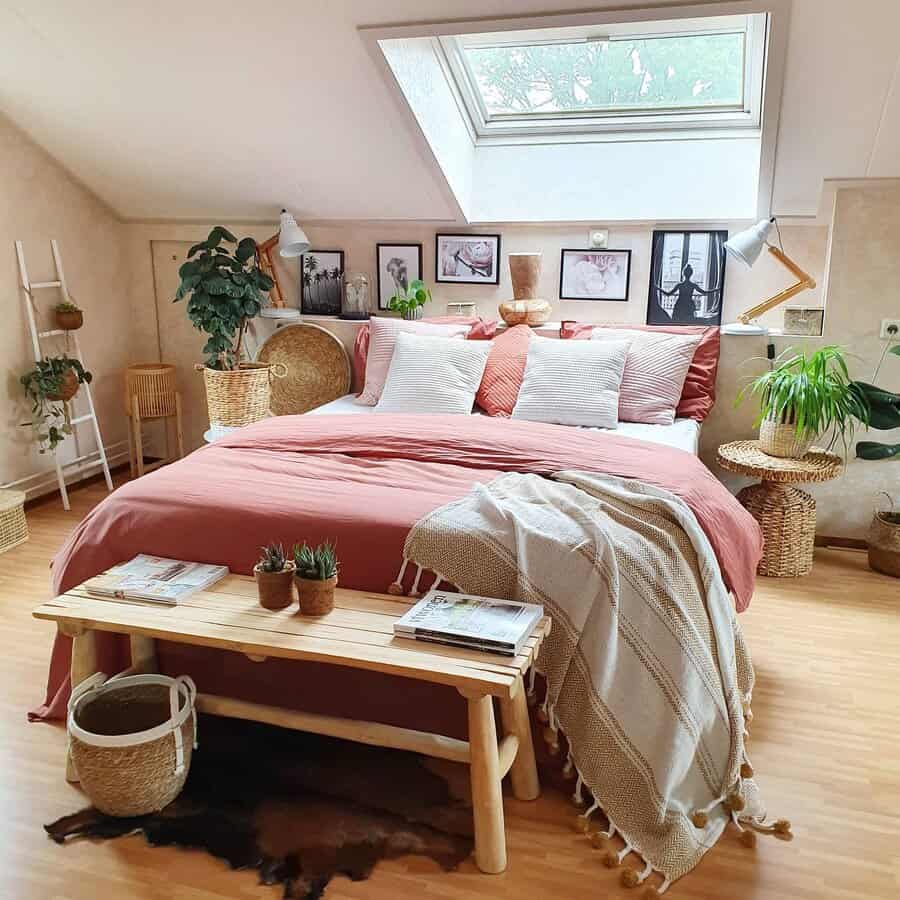 Boho bedroom with a skylight, pink bedding, and plants; decorative frames on walls, wooden bench at foot of bed, and woven accents