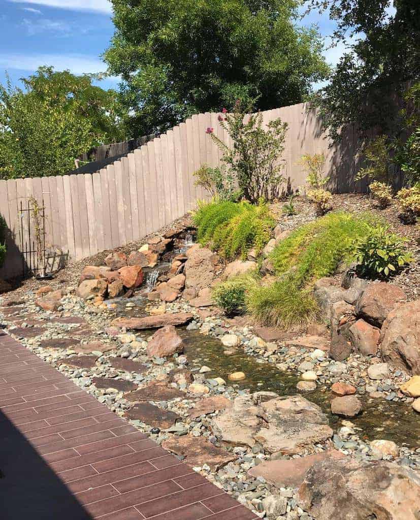 Backyard hardscaping design featuring a natural rock stream, wooden fence, greenery, and a tiled walkway