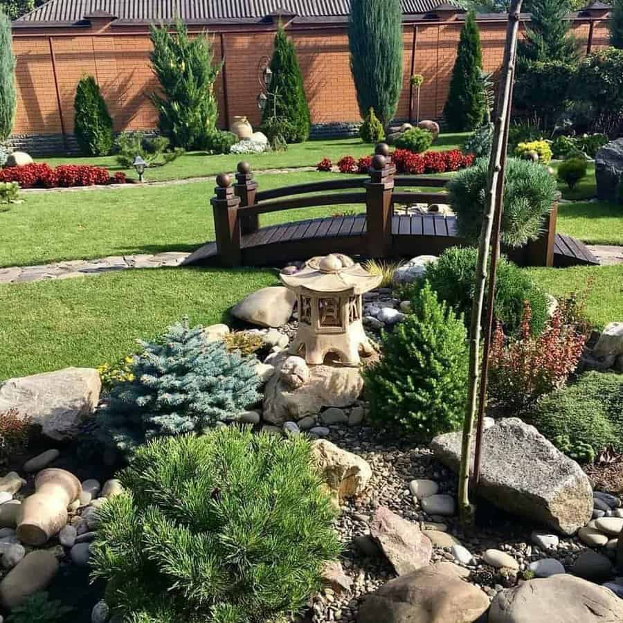 Japanese-inspired backyard hardscaping with a wooden bridge, stone lantern, decorative rocks, evergreens, and lush greenery