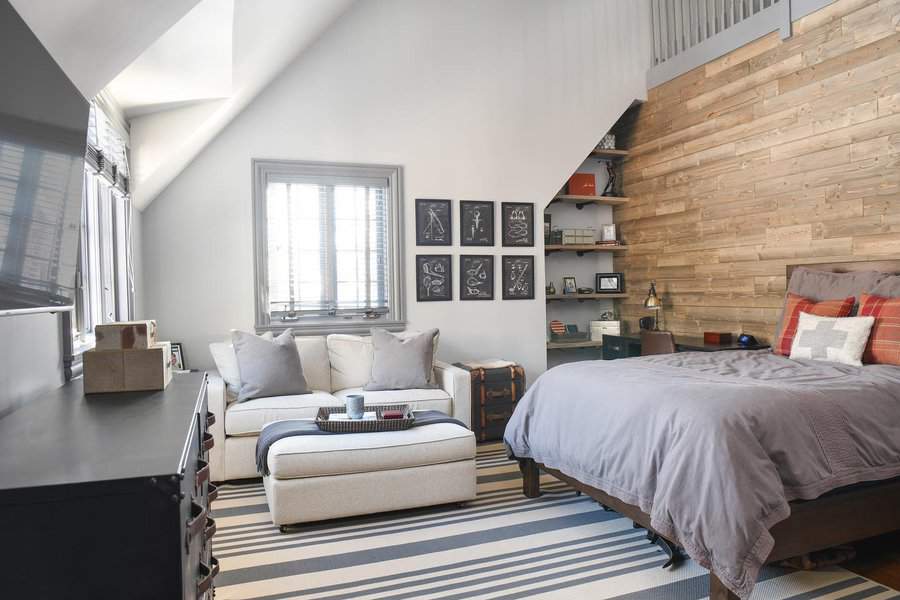 Modern attic bedroom with a rustic wood accent wall, neutral tones, and industrial decor. A cozy seating area and bold striped rug add style.