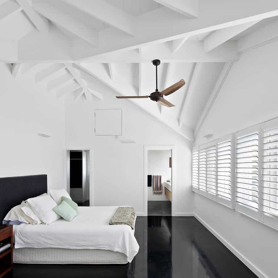 Minimalist attic bedroom with high vaulted ceilings, exposed beams, and sleek black floors. White walls and large windows create an airy feel.