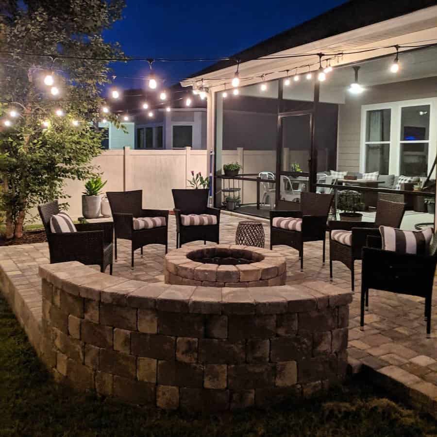 Cozy backyard with brick pavers, a fire pit, wicker seating, and string lights creating a warm and inviting outdoor gathering space