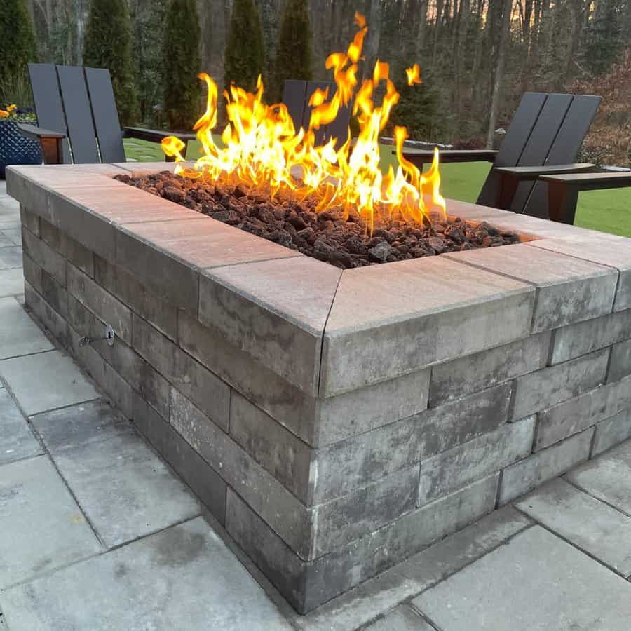 A stone fire pit with bright flames surrounded by patio chairs on a paved outdoor area with trees in the background