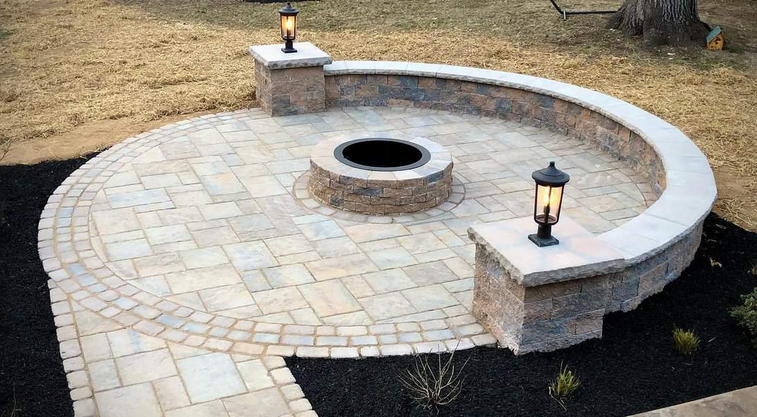 A circular stone patio with a fire pit in the center, surrounded by a low retaining wall with lanterns, grass and mulch on the sides