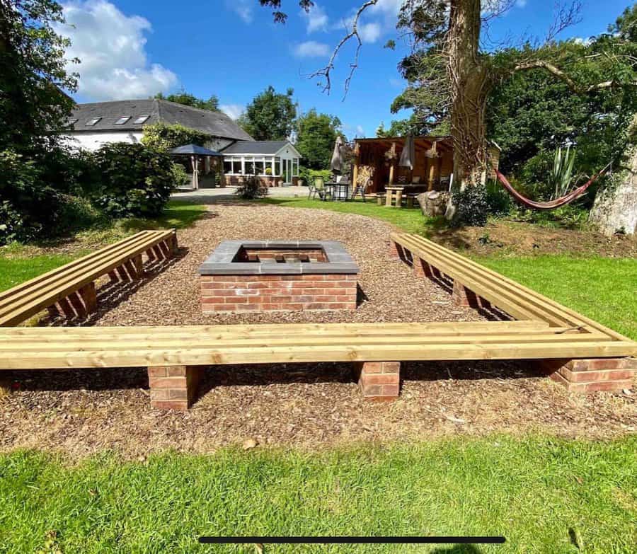 Backyard with wooden benches around a brick fire pit, a hammock to the right, and a house in the background under a blue sky