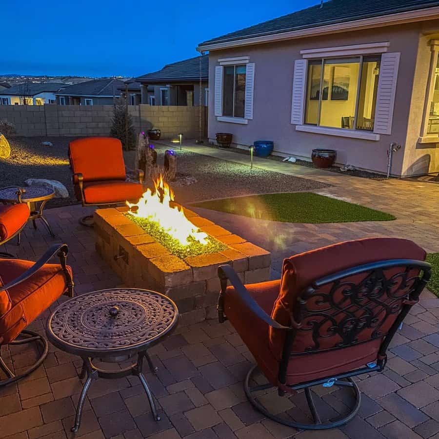 Backyard patio with red cushioned chairs around a lit fire pit on a brick surface, near a house at dusk