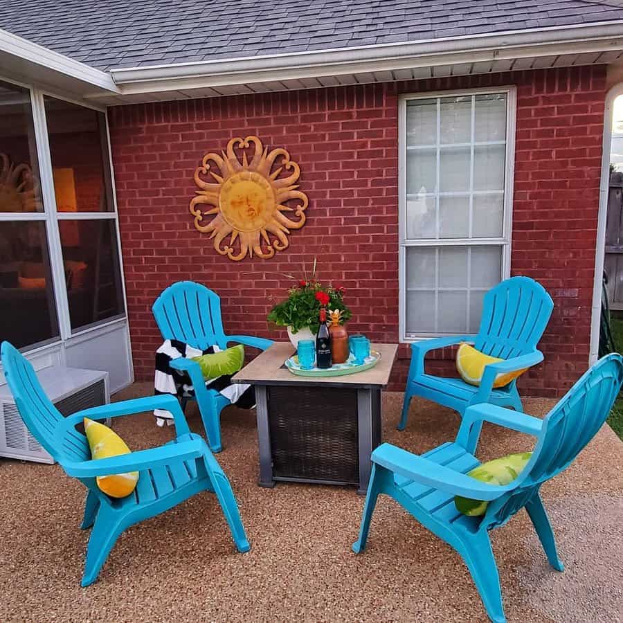 Patio with four turquoise chairs, a fire pit table, yellow pillows, and a metal sun decoration on a brick wall