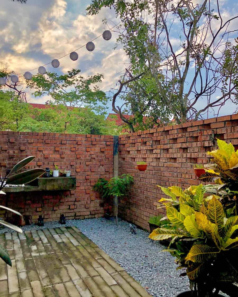 Rustic brick-walled patio with potted plants, string lanterns, gravel flooring, and a built-in outdoor shelf with greenery