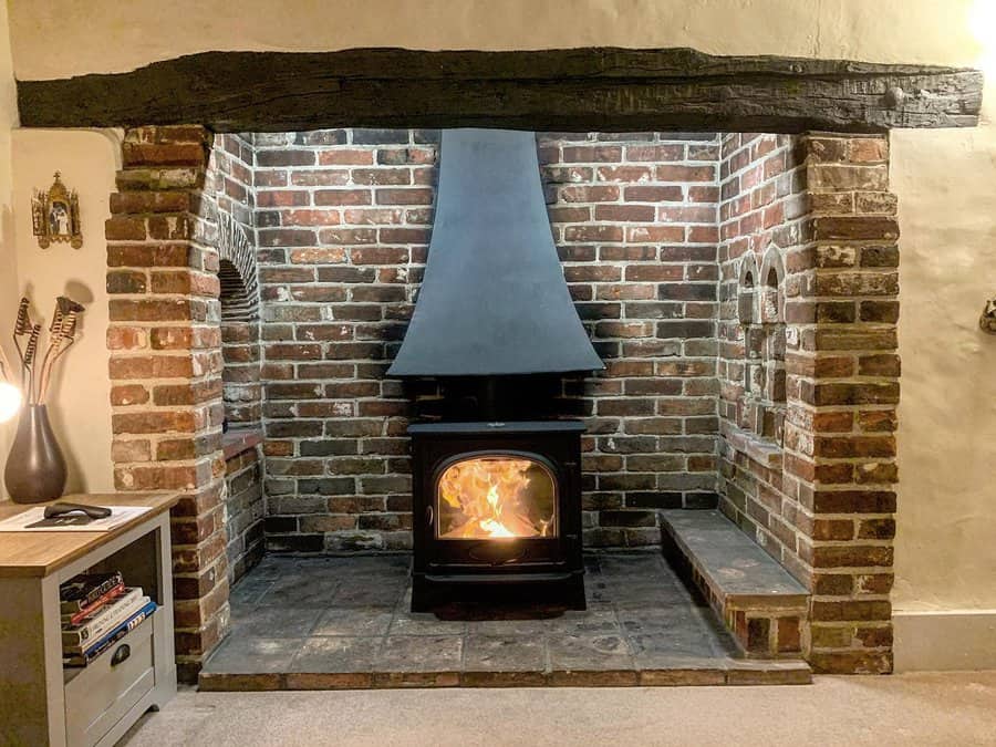 Cozy brick hearth with a wood stove and black metal hood, framed by rustic beams and built-in alcoves for a warm, traditional look.