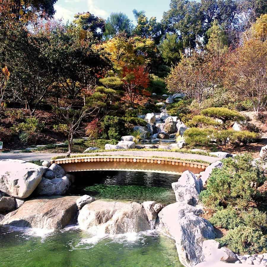 Serene Japanese garden with a wooden bridge over a clear pond, surrounded by lush greenery, cascading waterfall, and autumn foliage