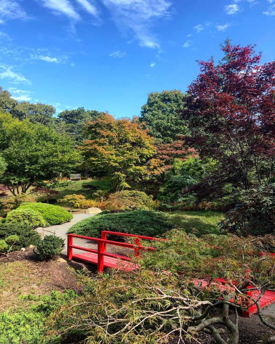 Vibrant Japanese garden with a striking red bridge, lush greenery, and colorful autumn foliage under a bright blue sky