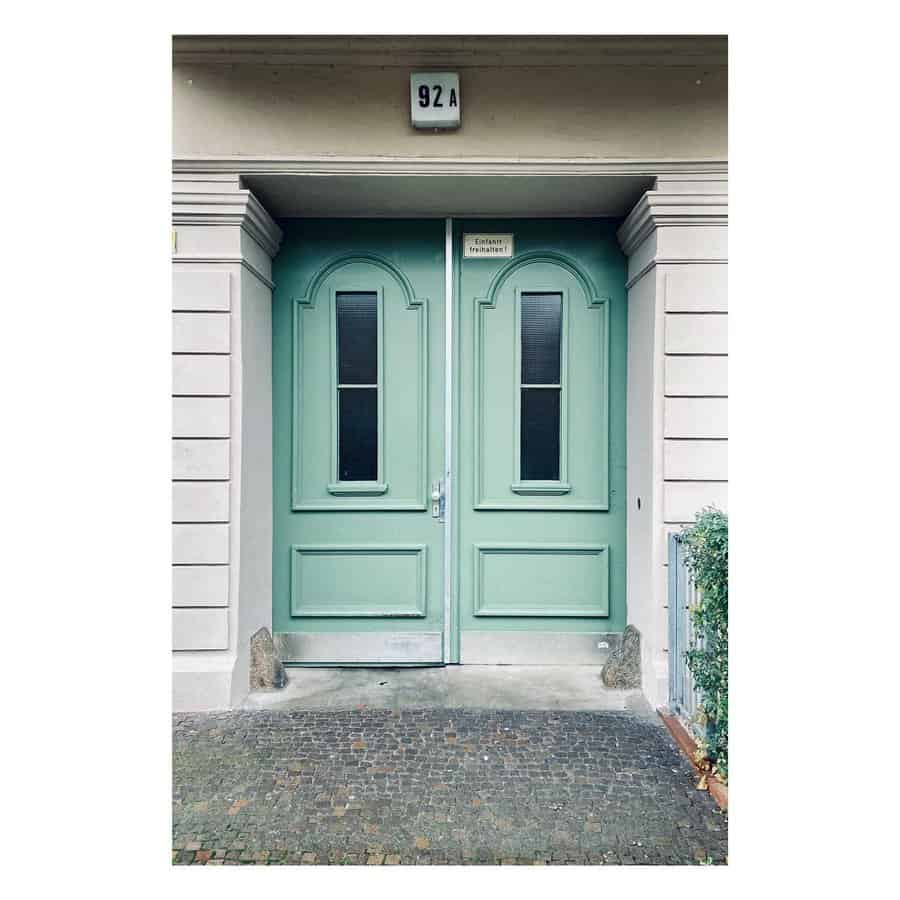 A mint green double door with black windows, set in a light gray building facade, with a small sign and house number 92A above it