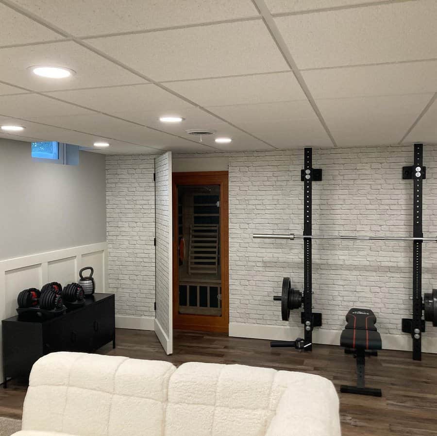 Multi-functional basement with a home gym, sauna, and cozy seating area, featuring a white brick accent wall and recessed lighting.