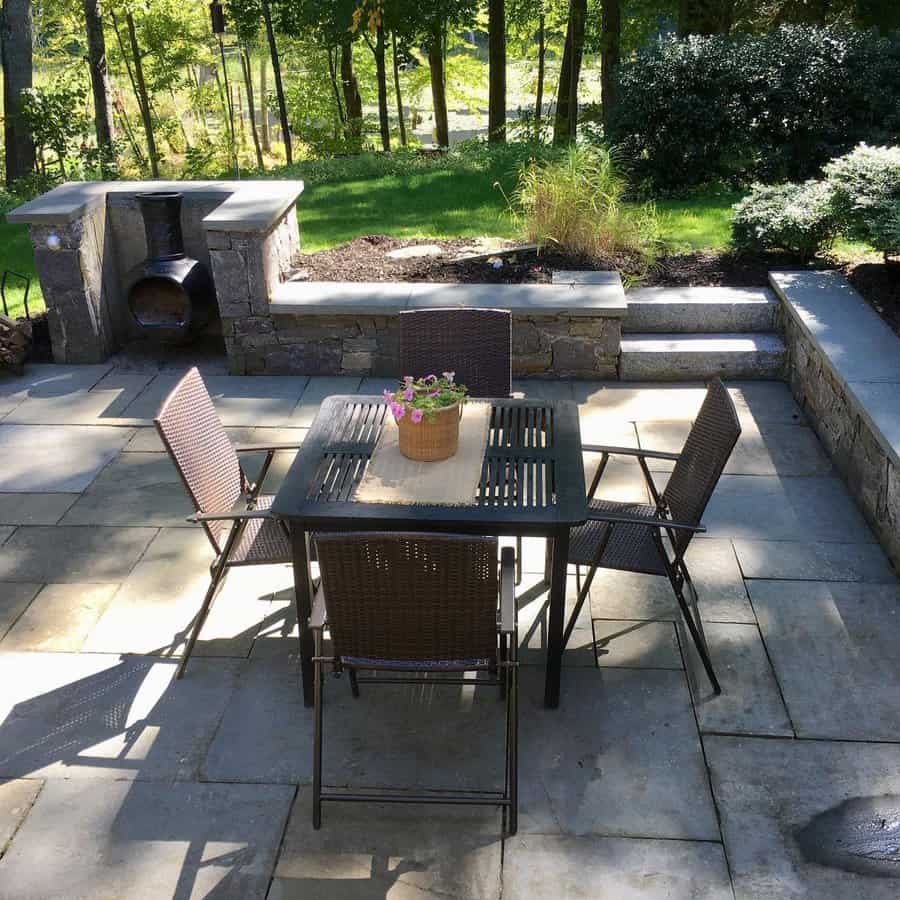 Outdoor patio with a metal table, four wicker chairs, and a potted plant centerpiece; stone chimney and lush greenery in the background