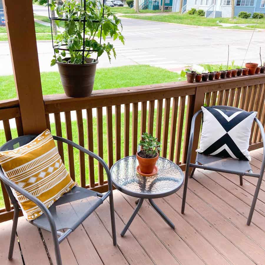 Two chairs with patterned cushions and a small table with a potted plant on a wooden porch, overlooking a grassy lawn and street