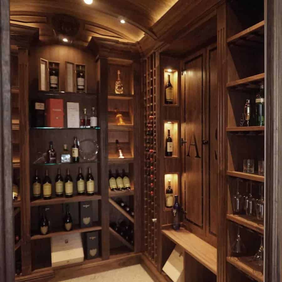 Wood-paneled wine cellar with wine bottles on shelves and racks, glasses on the right, and soft overhead lighting