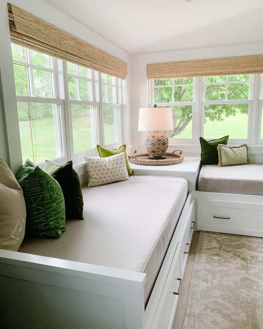 Cozy sunroom with twin daybeds, green and white pillows, a woven lamp on a side table, large windows, and greenery outside