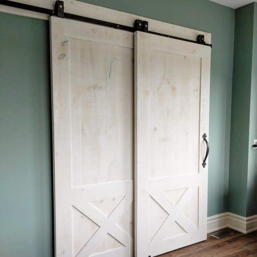 Two rustic white sliding barn doors on a green wall, featuring metal handles and hardware, with wooden flooring beneath