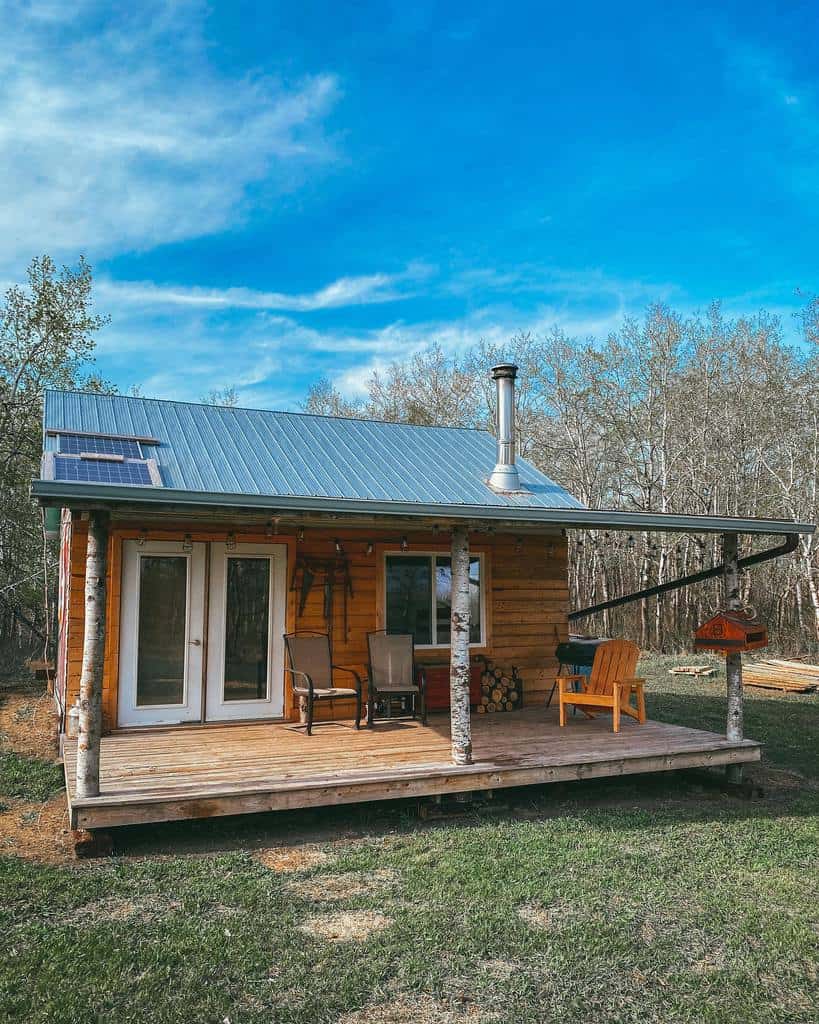 A small wooden cabin with a porch and chairs in a rural area under a blue sky