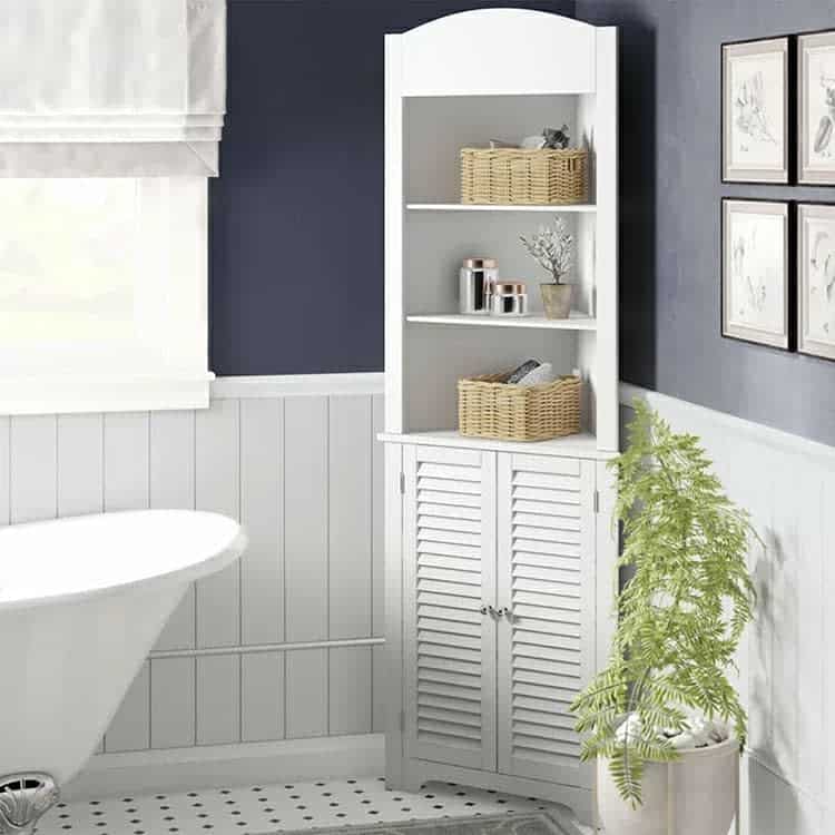 Corner bathroom cabinet with shutter doors and open shelves, holding baskets and toiletries, beside a fern and a bathtub