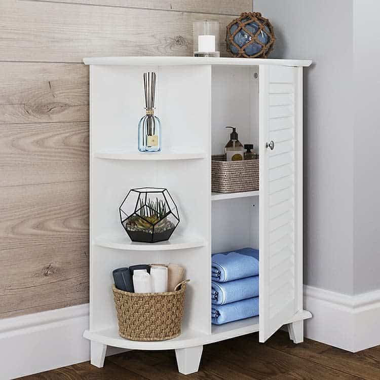 A white corner cabinet with open and closed shelves, holding towels, a candle, reed diffuser, small plant, and decorative items