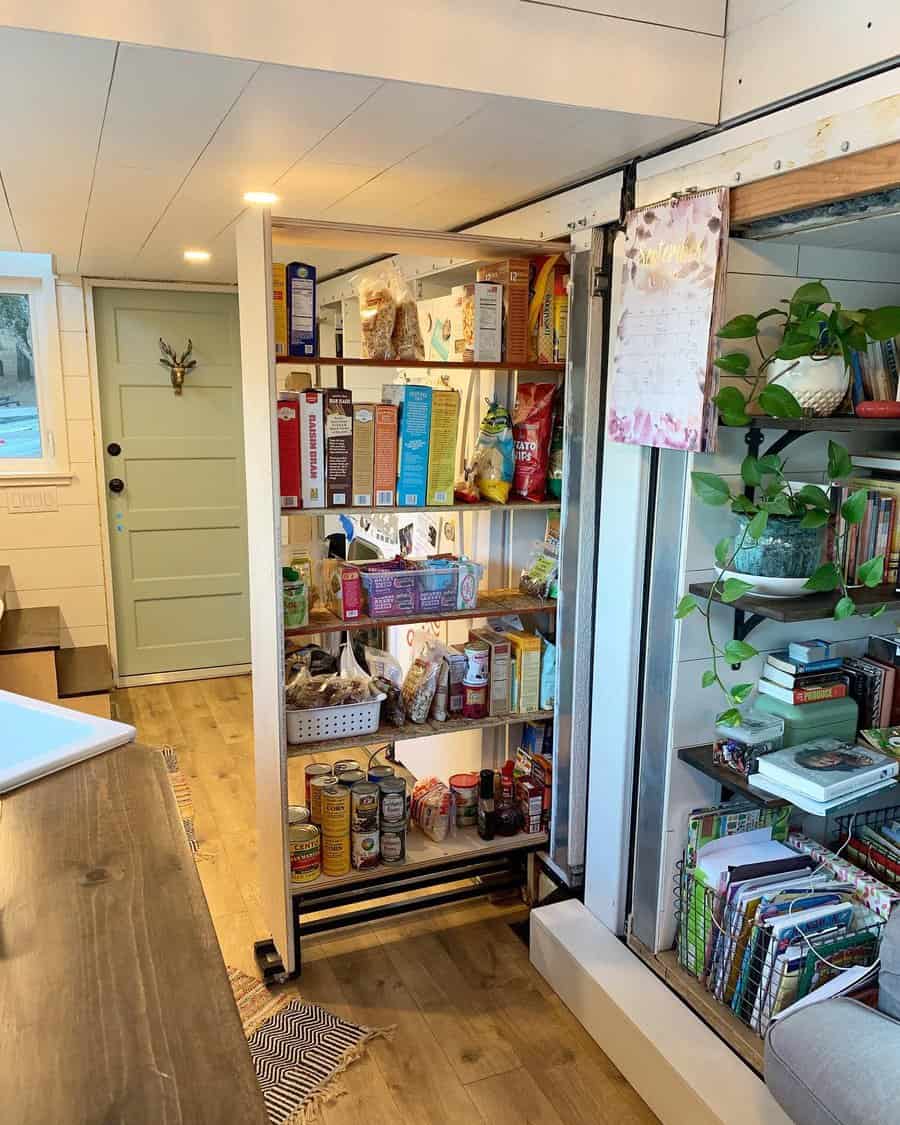 A pull-out pantry shelf filled with various food items, adjacent to a green door, and surrounded by plants and bookshelves