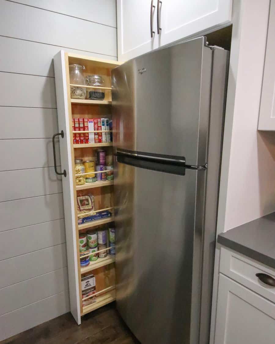 A tall, narrow pull-out pantry filled with jars, spices, and canned food is next to a stainless steel refrigerator in a modern kitchen