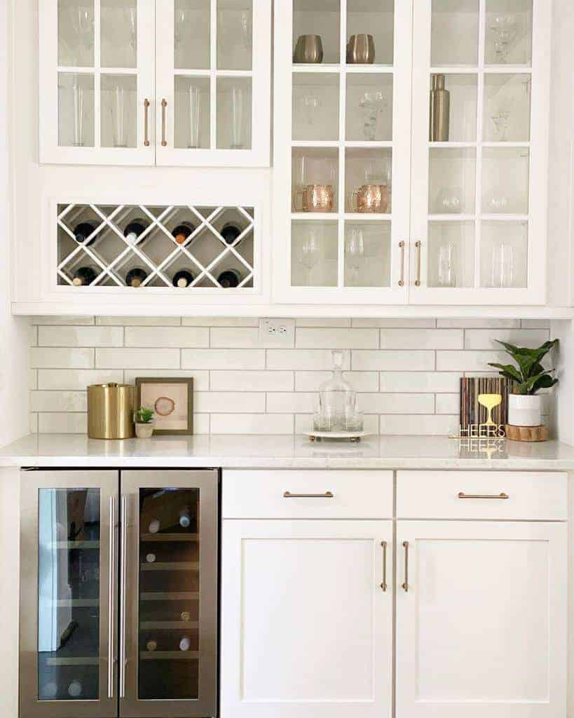 Elegant white kitchen with a built-in wine rack, glass cabinets, and a wine fridge, featuring gold accents and a subway tile backsplash