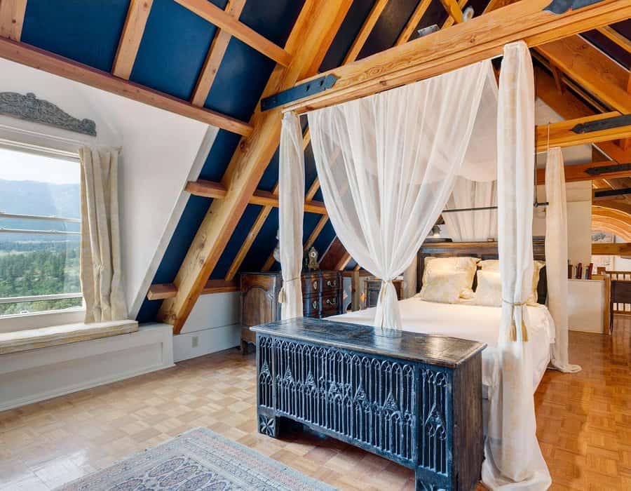 Rustic canopy bed with sheer drapes in a cozy attic bedroom featuring exposed wooden beams, a gothic-style chest, and scenic mountain views