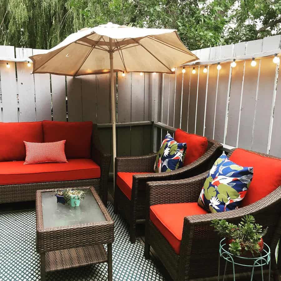 Patio with red-cushioned wicker furniture, colorful pillows, a beige umbrella, string lights, and a small glass-top table with plants