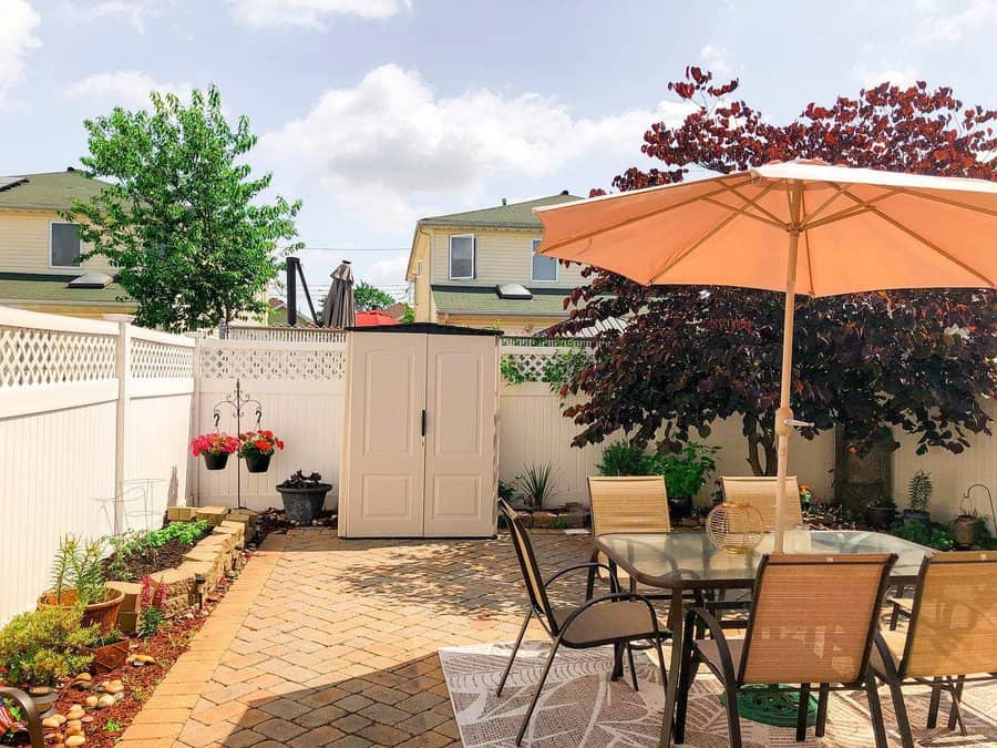 Sunny patio with table, chairs, and umbrella; potted plants and storage shed against white fence; trees and houses in the background