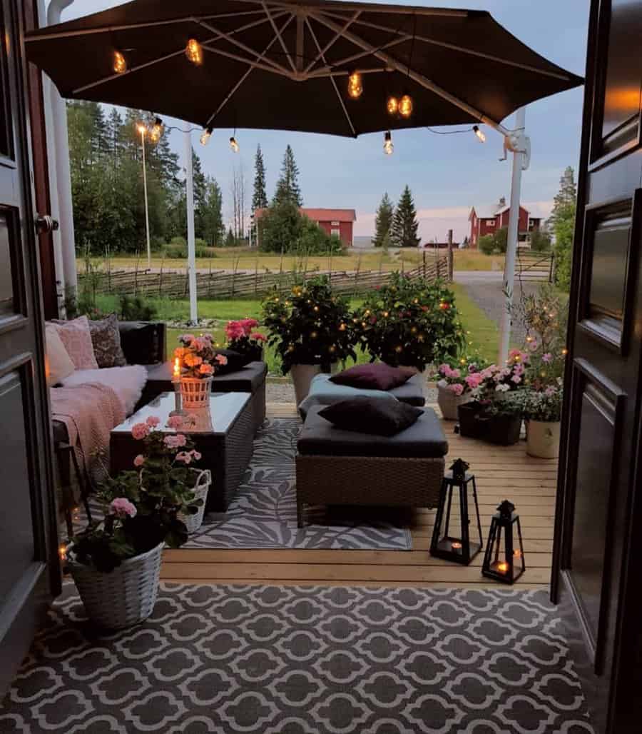 Cozy outdoor patio with a wicker sofa, cushions, plants, lanterns, and a large umbrella; scenic countryside view in the background