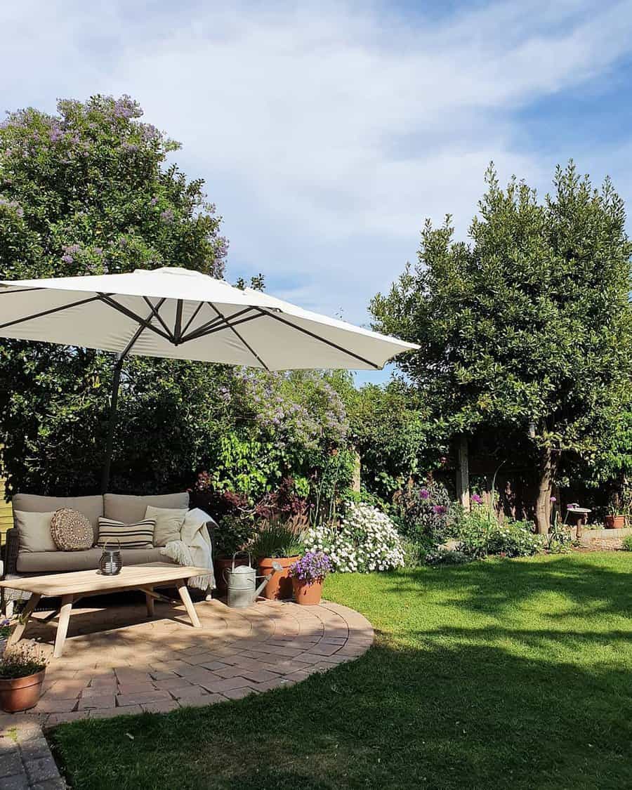 A garden with a seating area under a large white umbrella, surrounded by trees and flowers, on a sunny day