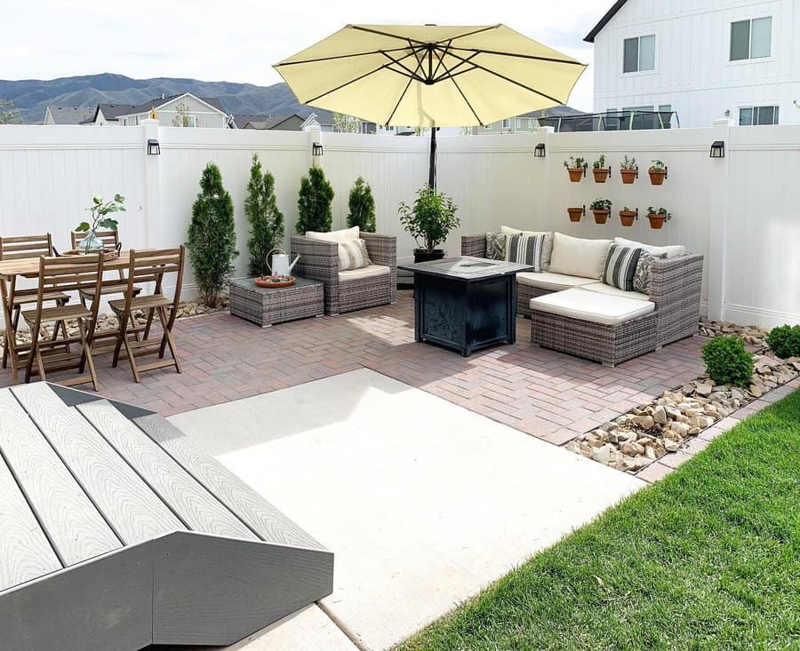 Patio with wicker furniture, table, umbrella, and wall planters on a brick area, surrounded by a white fence and greenery