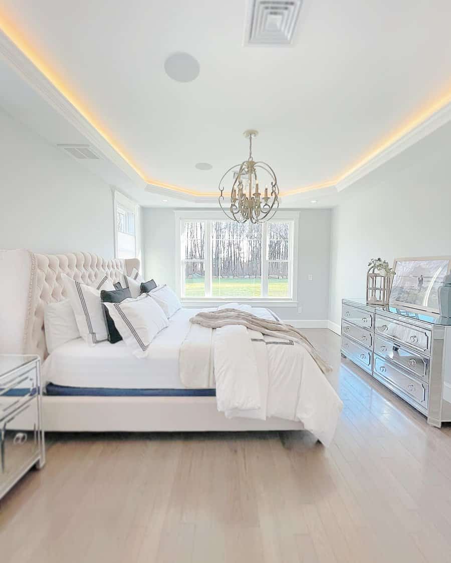 Bright bedroom with a tufted bed, chandelier, mirrored dresser, and large window overlooking a wooded area in soft, neutral tones