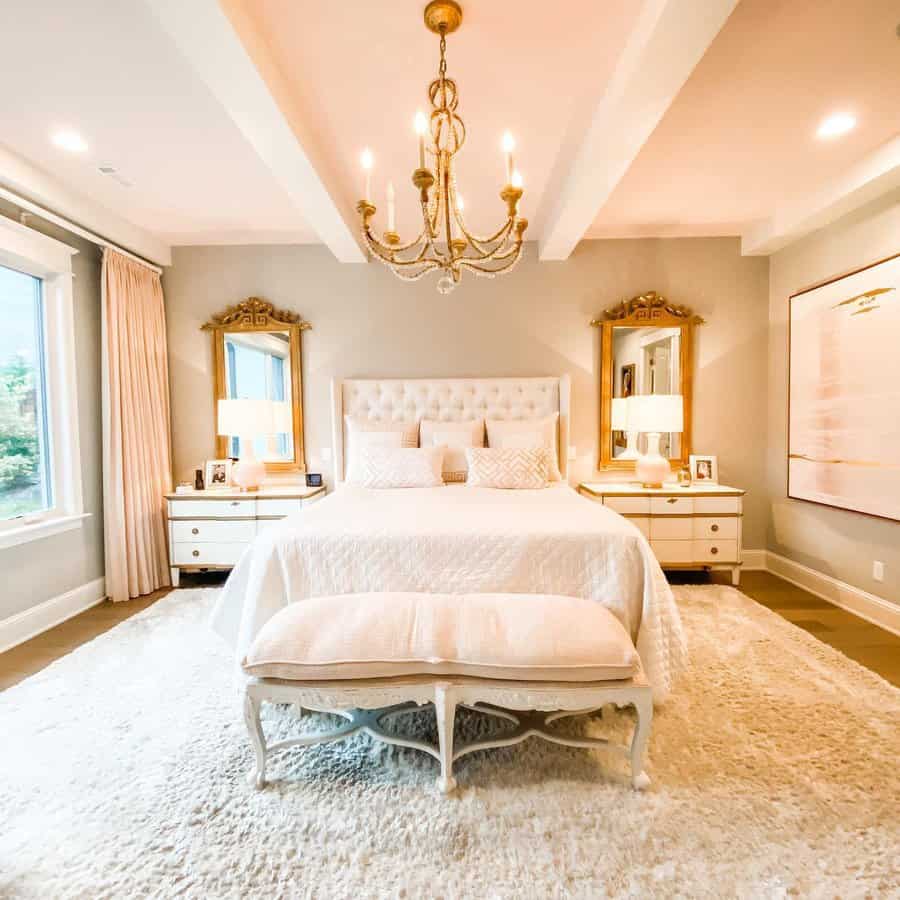 Elegant master bedroom with a large bed, tufted headboard, chandelier, and mirrors; beige and white color scheme with soft lighting