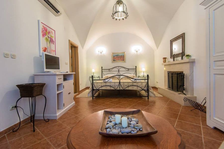 Master bedroom with a wrought iron bed, TV on a shelf, wicker chair, and decorative stones on a table; warm lighting and wall art