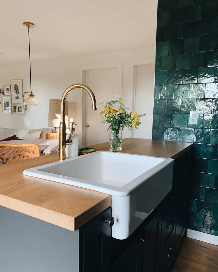 Modern kitchen with a white farmhouse ceramic sink, brass faucet, wooden countertop, and green tiled wall, vase with flowers on the counter