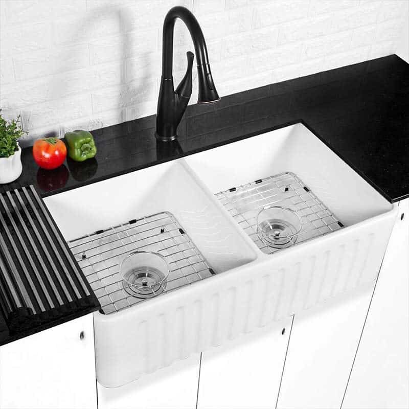 A double white kitchen ceramic sink with a black faucet, metal grid racks, and vegetables on the counter: tomato, cucumber, and bell pepper