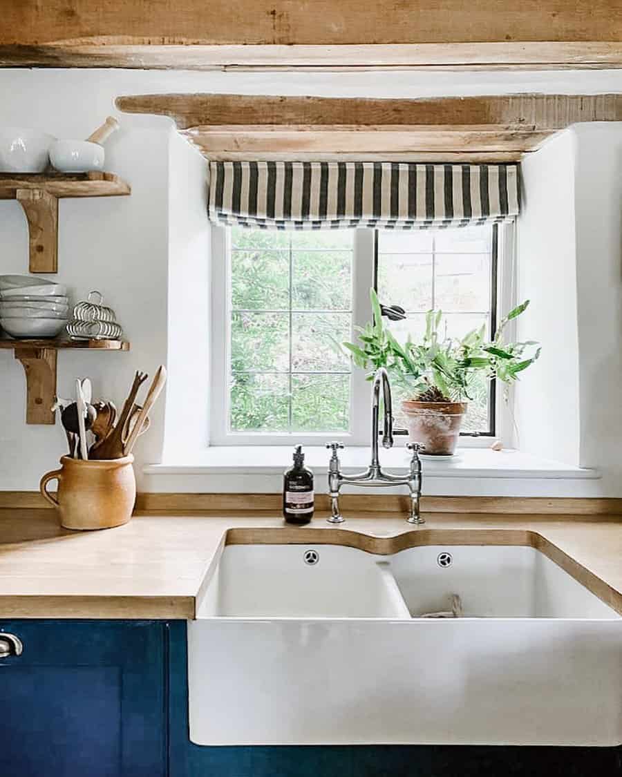 Rustic kitchen with a farmhouse ceramic sink, wooden counter, striped window shade, potted plant, and shelves with dishes and utensils