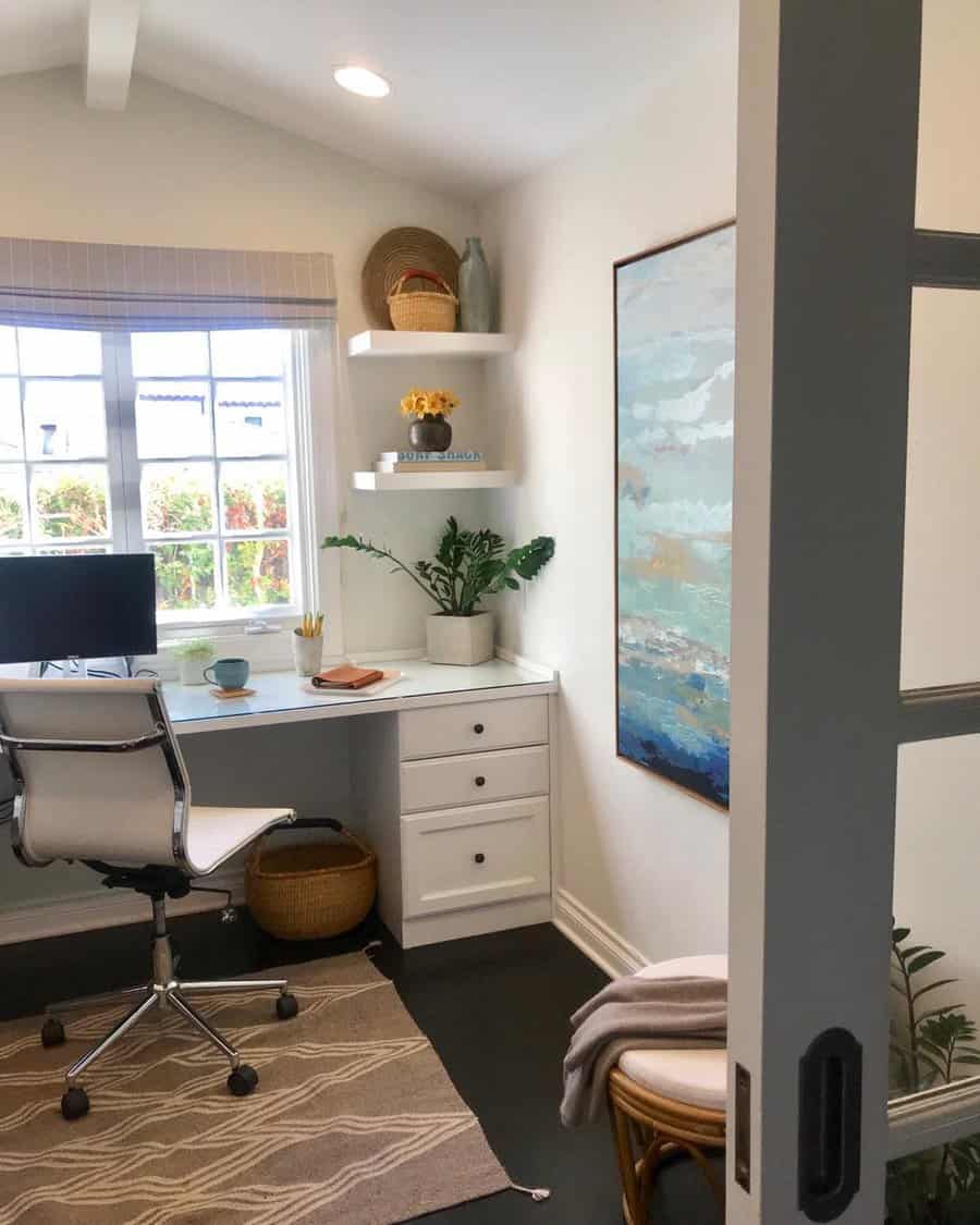 A cozy home office with a desk, white chair, and monitor; shelves holding plants and decor; a large bay window letting in natural light