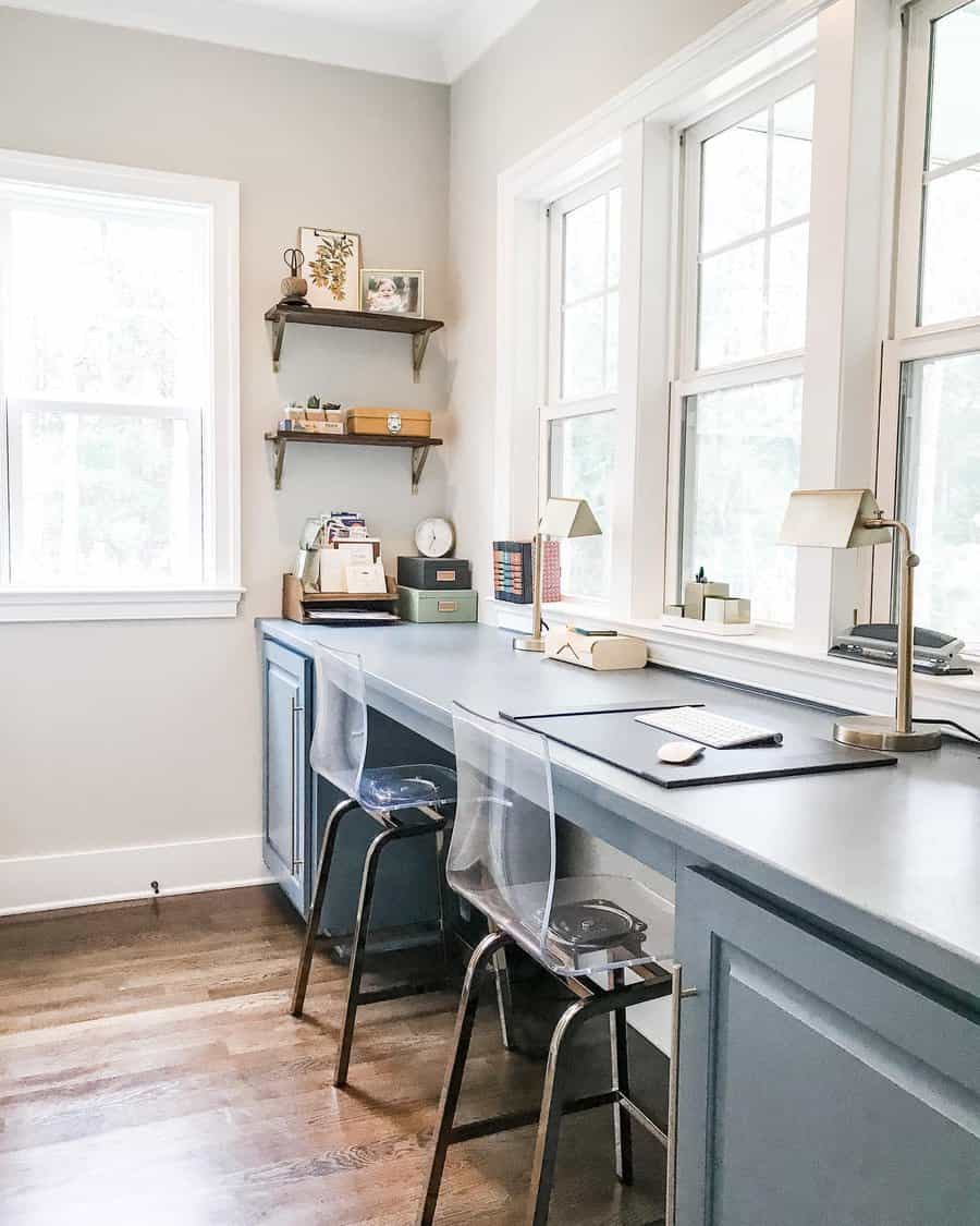 A bright home office with two transparent chairs, blue desk, lamps, and wall shelves, set against large bay windows